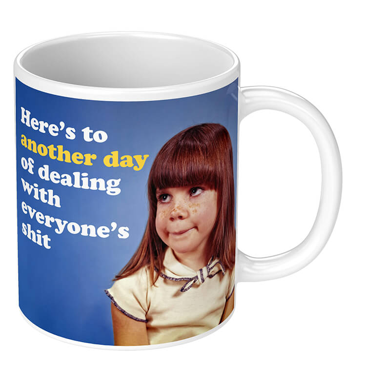 A novelty mug with a retro picture of an a bored looking girl looking into the middle distance. White rounded text above her reads Here's to another day of dealing with everyone's shit